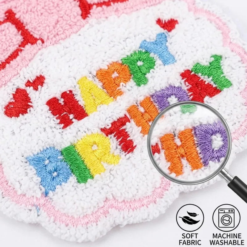 A close-up of a white textured blanket with colorful embroidered 'HAPPY BIRTHDAY' lettering and a pink border, shown with a magnifying glass over the letters and small icons indicating soft fabric and machine washable.