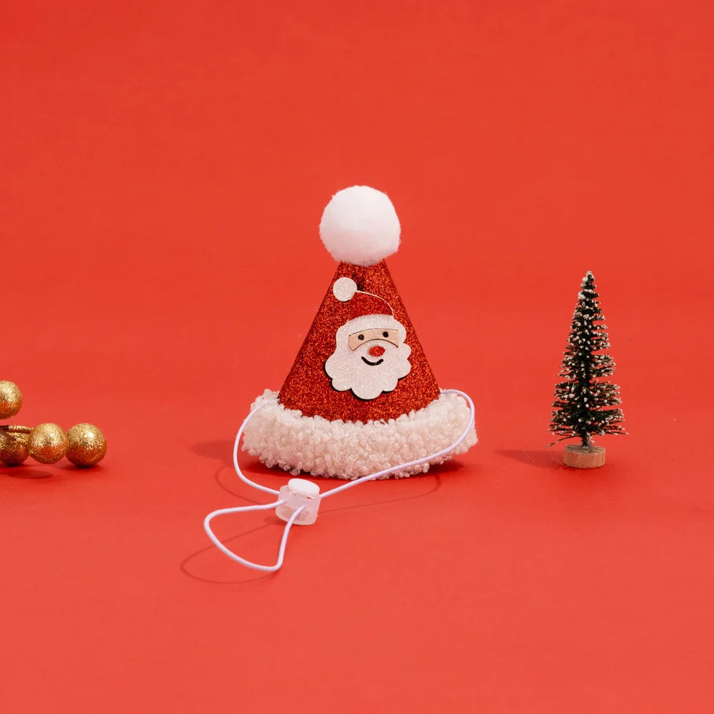 A festive red glitter Santa hat with a white faux-fur trim, a white pom-pom, and a printed Santa face. It is shown on a bright red background with a white ear-loop strap, styled with a miniature Christmas tree and gold beads.