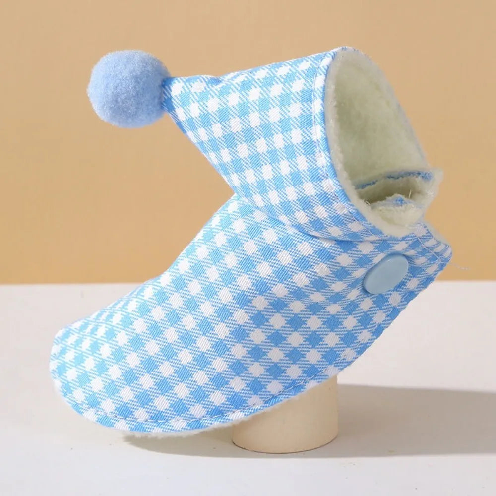 A baby bootie with a light blue and white gingham pattern, featuring a soft blue pom-pom on the pointed toe and a cream-colored fleece lining, displayed on a small stand.