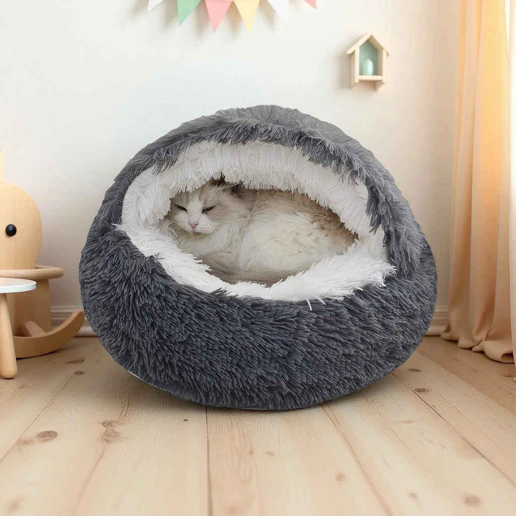 A fluffy round pet bed in dark gray and white, with a white cat resting inside, set on a light wood floor in a cozy room.