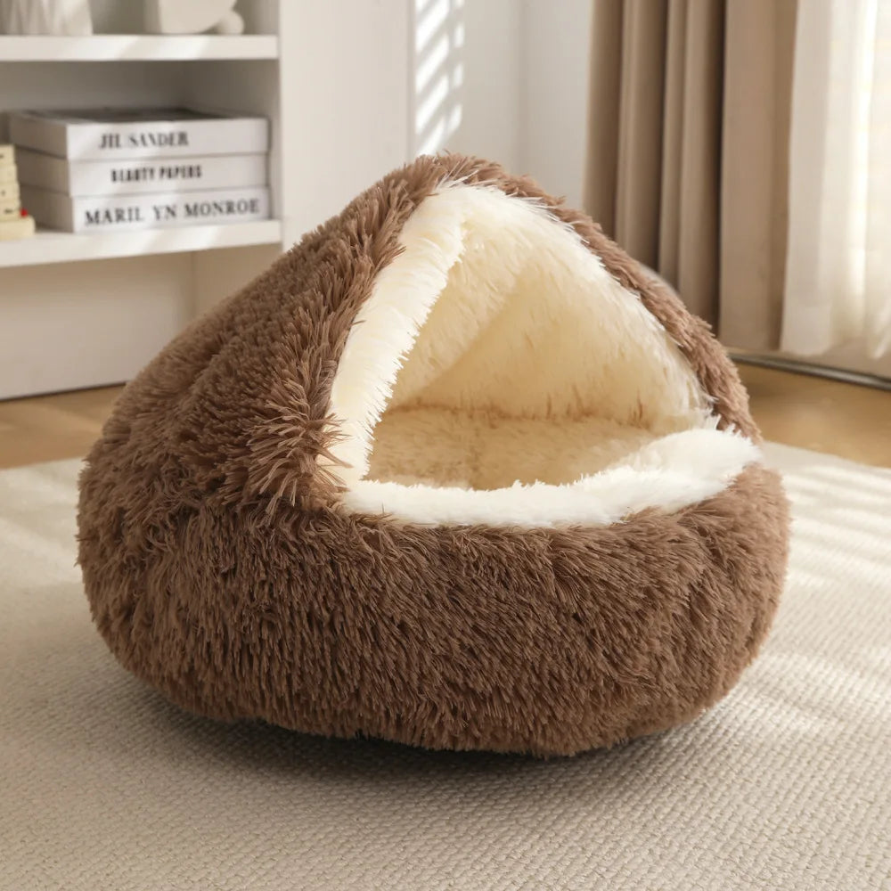 A brown plush pet bed with a cream sherpa-lined opening, placed on a light rug in a softly lit room with a bookshelf and curtains in the background.