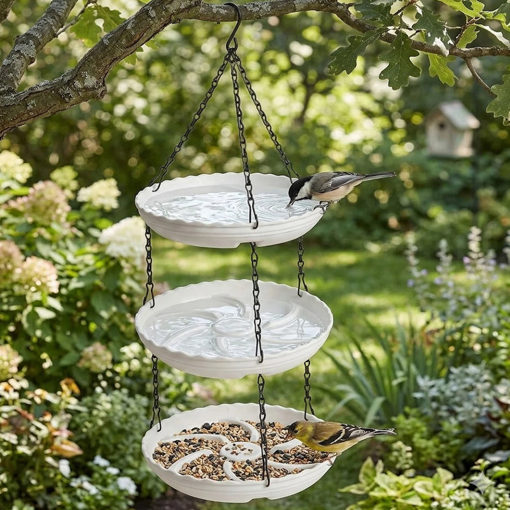 A three-tier hanging bird feeder with black chains and white scalloped bowls, shown outdoors in a garden with two birds perched on the bowls.