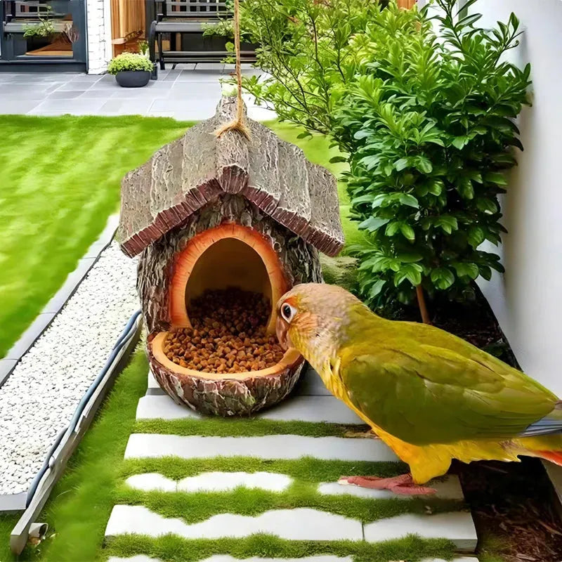 A small green-and-yellow parrot stands beside a log-shaped bird feeder filled with kibble in a landscaped garden with grass, plants, and stone steps.