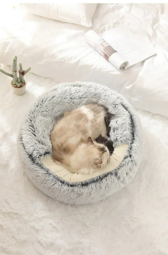 A fluffy round pet bed with a gray faux-fur exterior and cream interior, shown on a white bed with a cat curled up inside. A small cactus and rolled magazine are nearby.