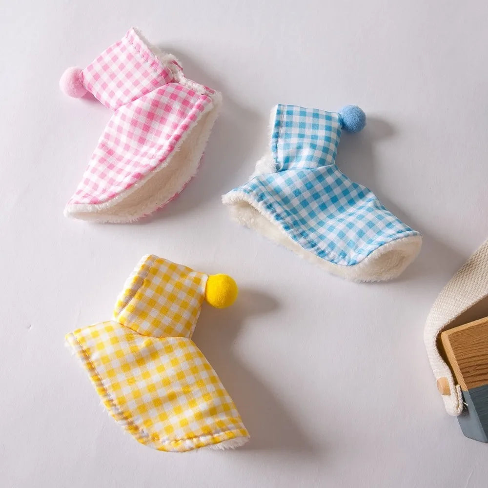 Three small hooded fabric pouches or pouches with gingham checks are arranged on a light surface. One is pink and white, one is blue and white, and one is yellow and white, each with a small pom-pom accent at the hood.