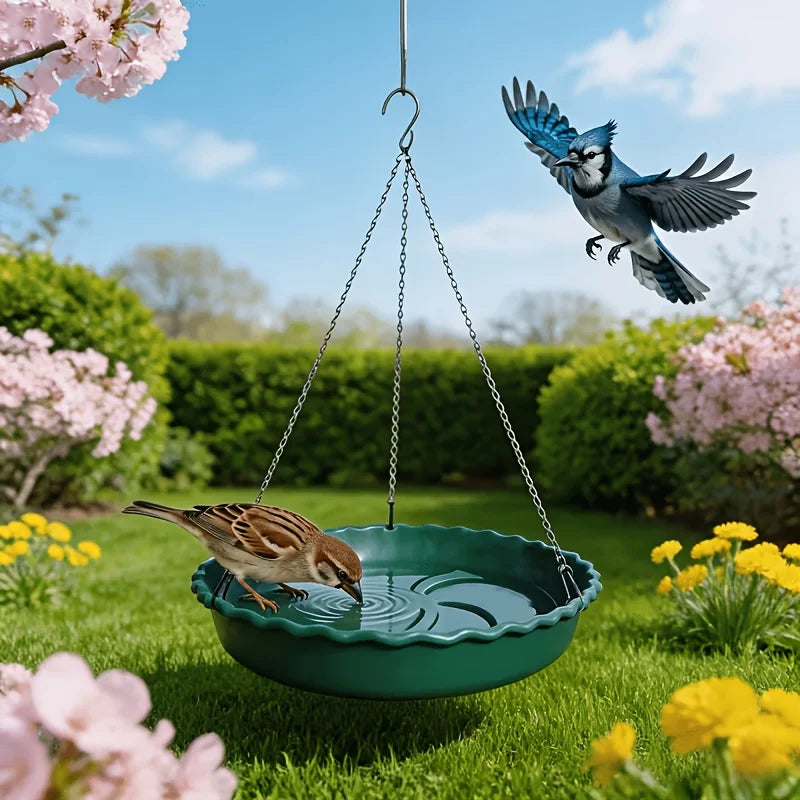 A green hanging bird bath with two birds in a sunny garden. One bird is perched on the rim while the other drinks from the water in the bowl.