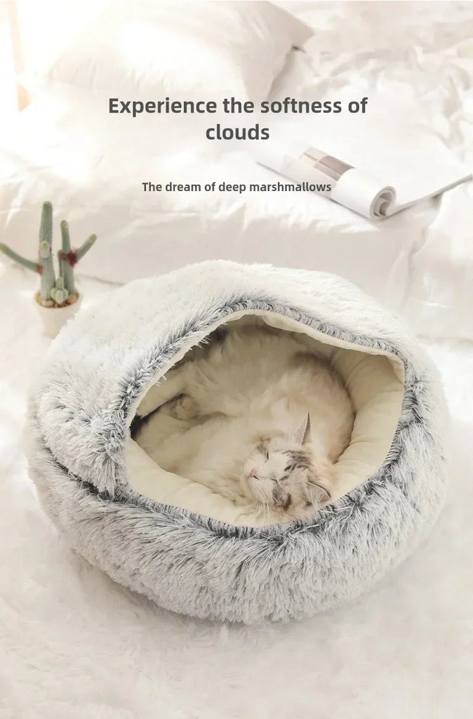 A fluffy round pet bed with a cat curled up inside, placed on a soft white blanket with pillows and a small plant in the background. Text on the image reads, 'Experience the softness of clouds' and 'The dream of deep marshmallows.