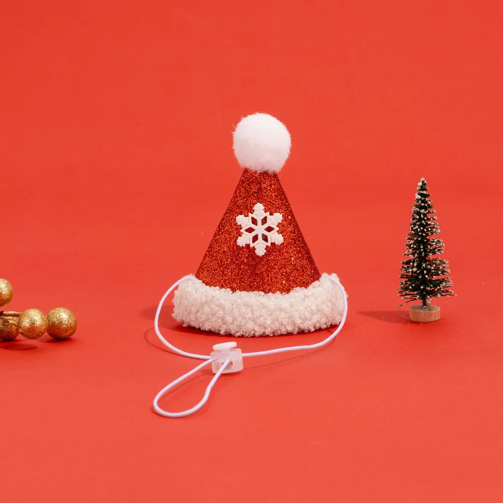 A miniature red glitter Santa hat with a white faux-fur trim, a white pom-pom, and a white snowflake ornament on the front, attached to a white elastic cord. It is displayed on a solid red background with decorative props, including a small Christmas tree and gold beads.
