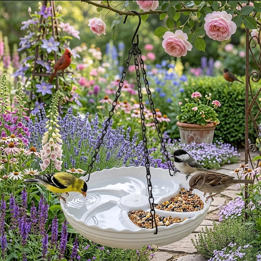 A white ceramic hanging bird bath with a divided tray, shown in a lush garden with birds feeding and drinking. The bird bath is suspended by black chains from a tree branch, surrounded by pink roses, purple flowers, and several birds.