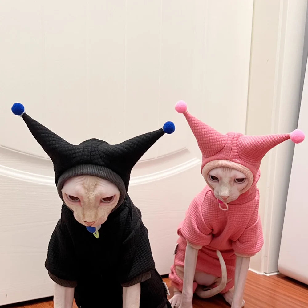 Two hairless cats wearing jester-style pet costumes, one in black with blue pom-poms and one in pink with pink pom-poms, posed indoors in front of a white door.