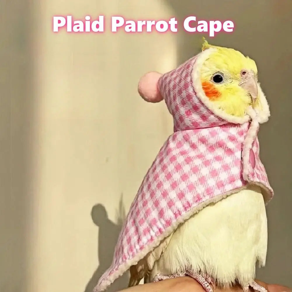 A small yellow parrot wearing a pink and white plaid hooded cape, perched on a person's hand against a beige wall with soft sunlight and shadows.