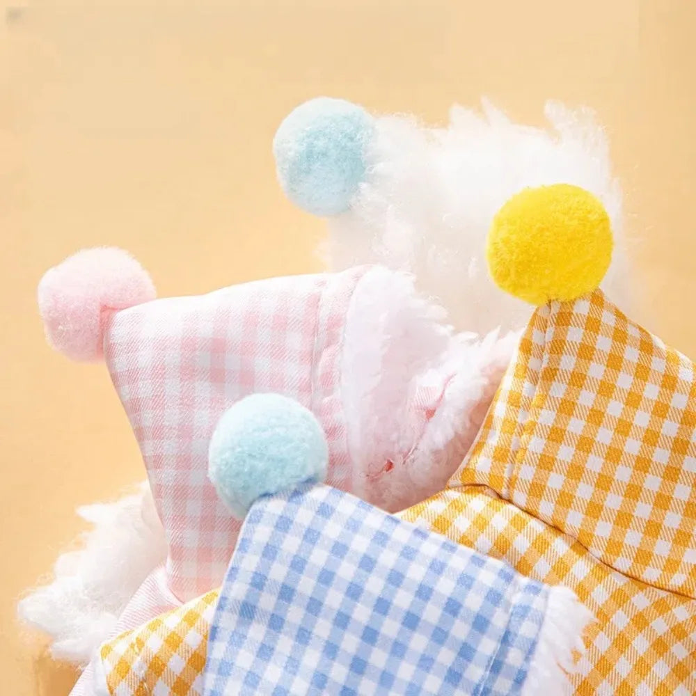 Three small plush toy hats or soft hats with gingham checks in pink, yellow, and blue, each topped with a fuzzy pom-pom in pastel pink, yellow, and blue, arranged against a warm beige background.