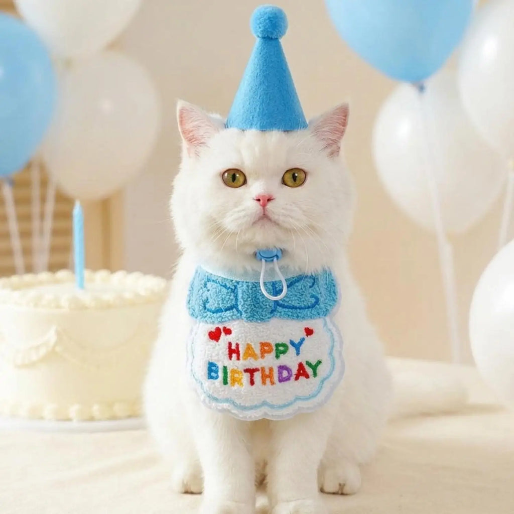 A white cat wearing a blue party hat and a blue bow-tie bib with colorful 'HAPPY BIRTHDAY' lettering, posed in front of a birthday cake and balloons.