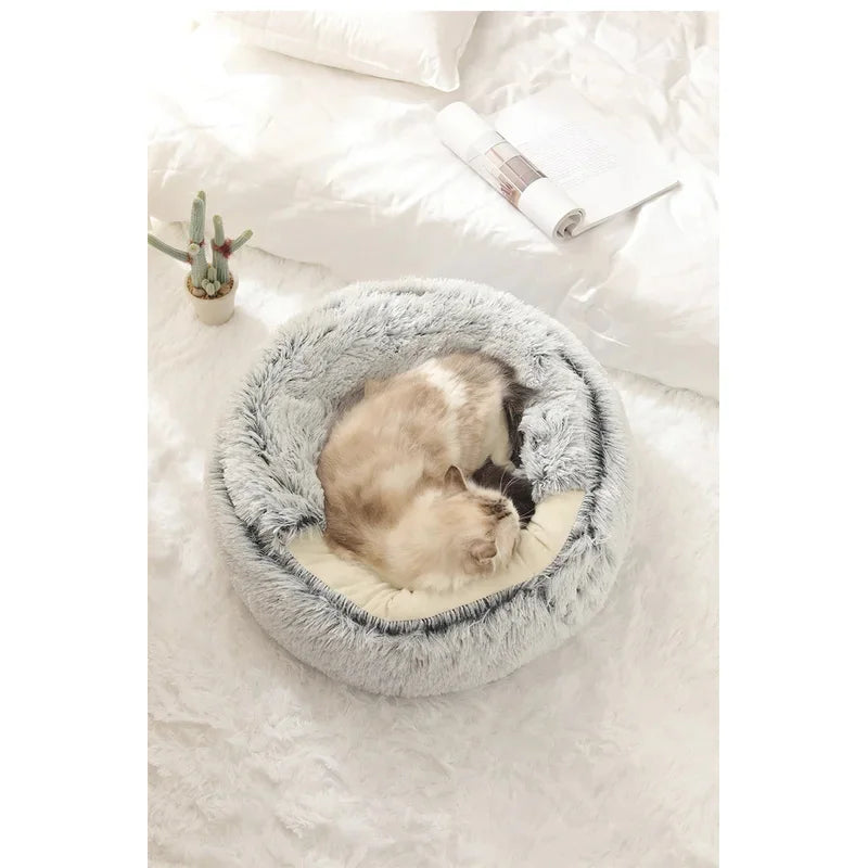 A fluffy round pet bed in light gray and cream, with a cat curled up inside, placed on a white bed with a small potted cactus nearby.
