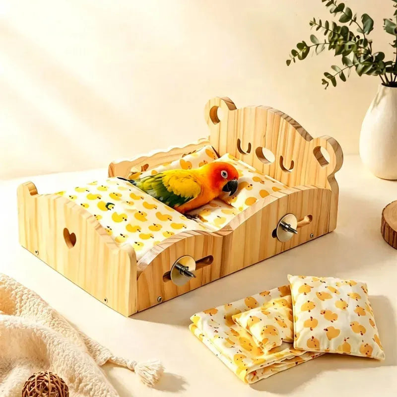 A wooden pet bed with a bear face headboard and metal ball toys, shown with a small parrot inside. The bed is dressed in a yellow and white duck-print fabric, and matching folded sheets and pillows are placed beside it on a light beige surface.
