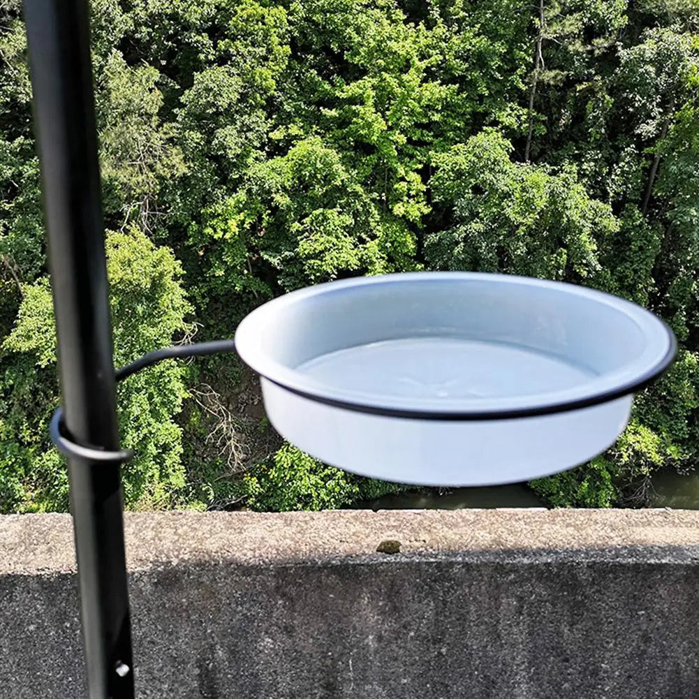 White metal bird bath with black rim on a metal stand against a green forest background.