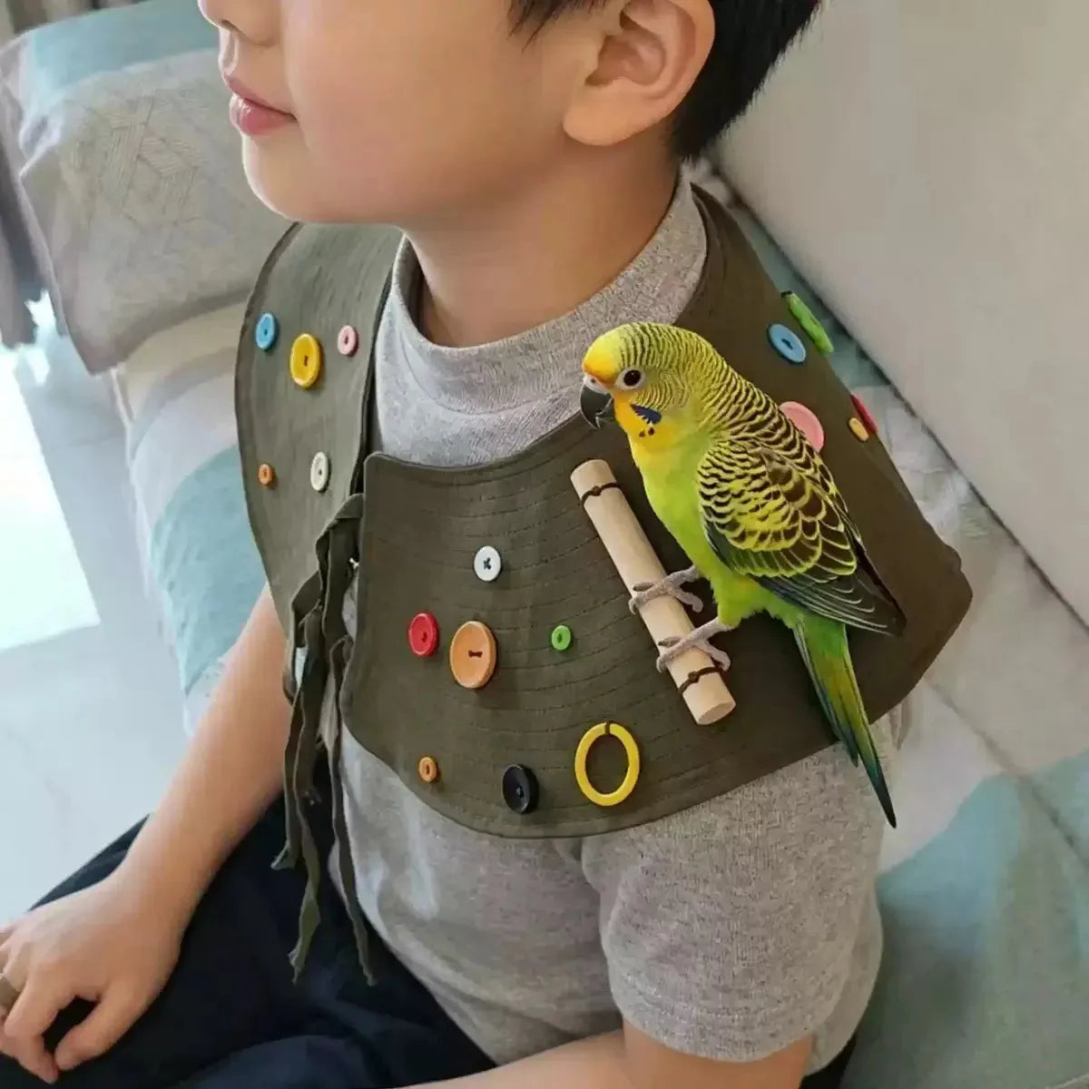 A child wearing a brown fabric bib decorated with colorful buttons and a small wooden toy, with a green and yellow budgerigar perched on the bib, in a home setting.