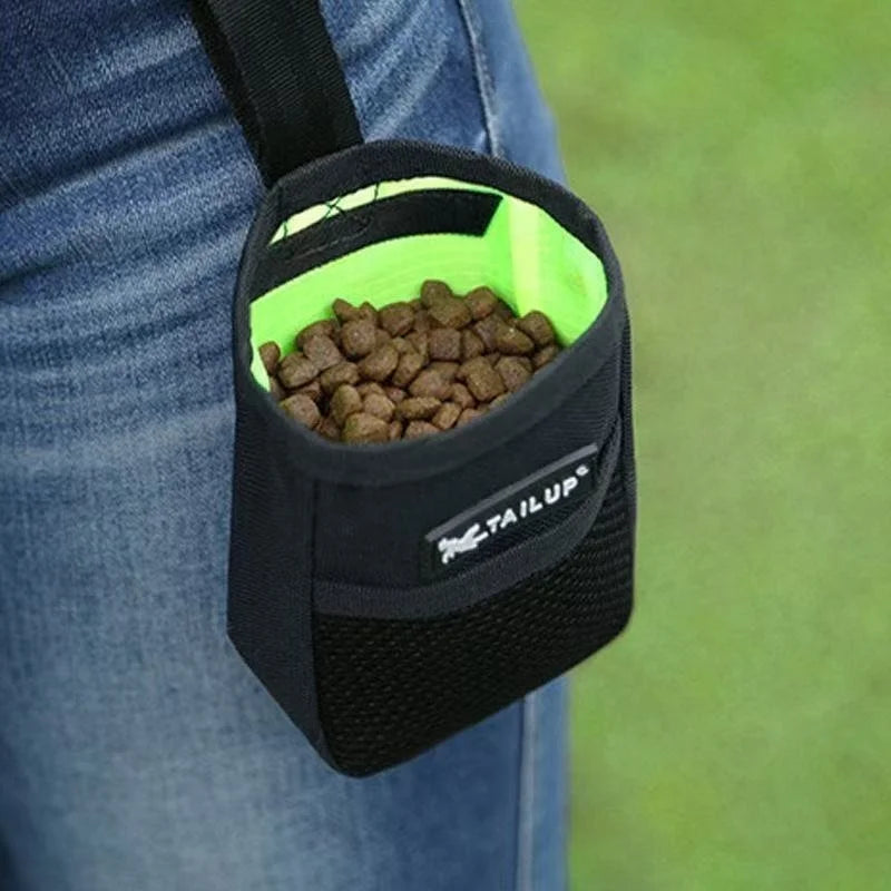 Black and green pet treat pouch filled with kibble held by a person wearing blue jeans.